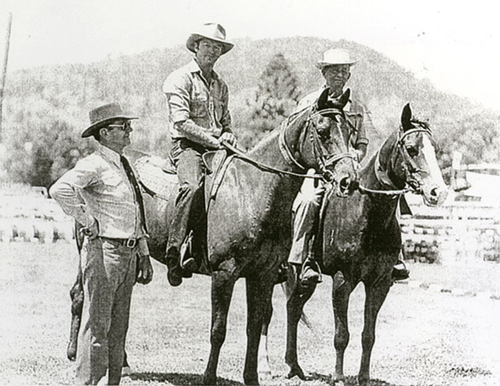 An old, grainy black and white photo of two men on horses and another man standing