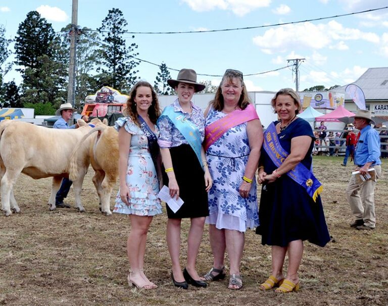 Four women wearing sashes standing on showgrounds in front of some cows